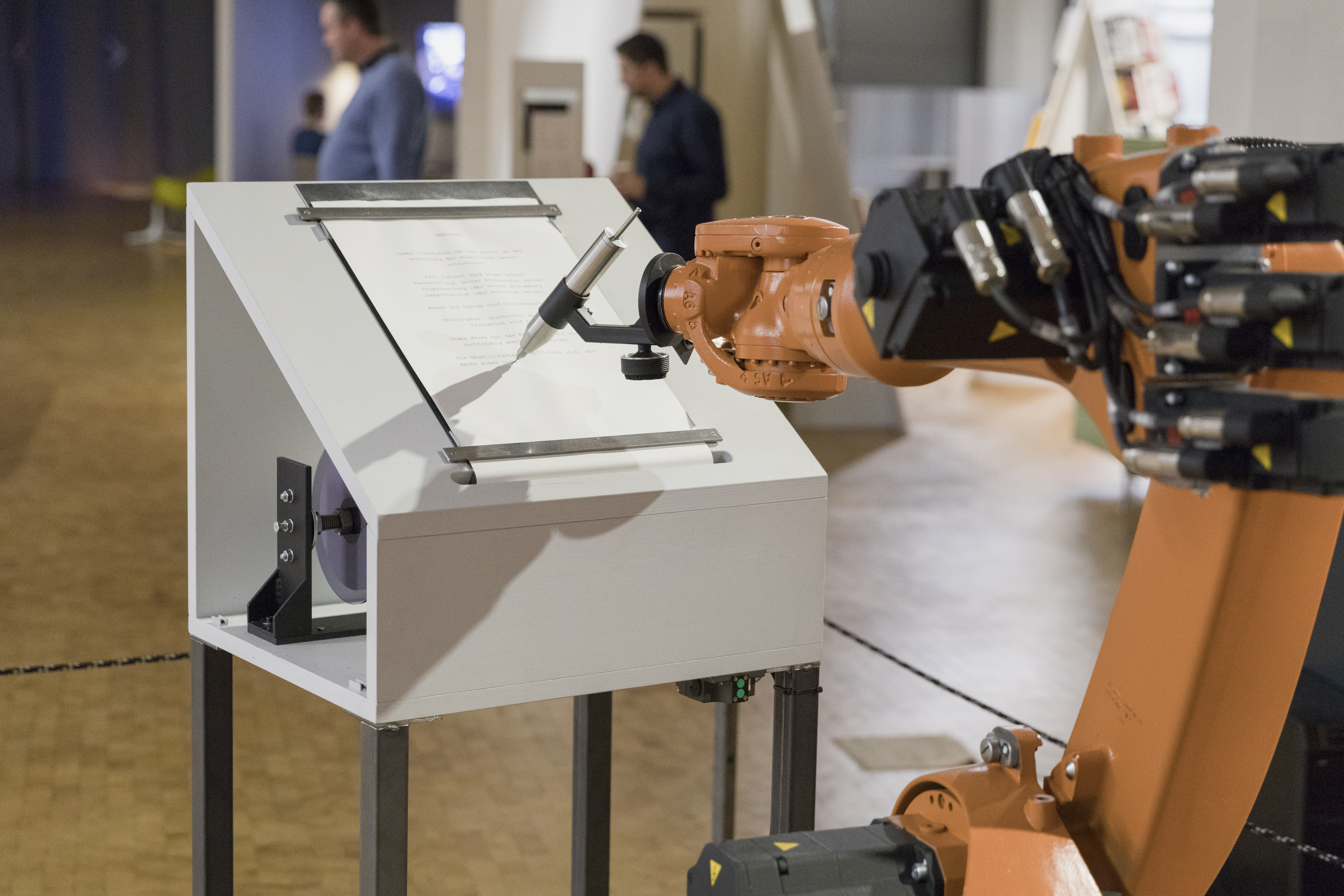 An orange robot arm writes with a large silver pen on paper