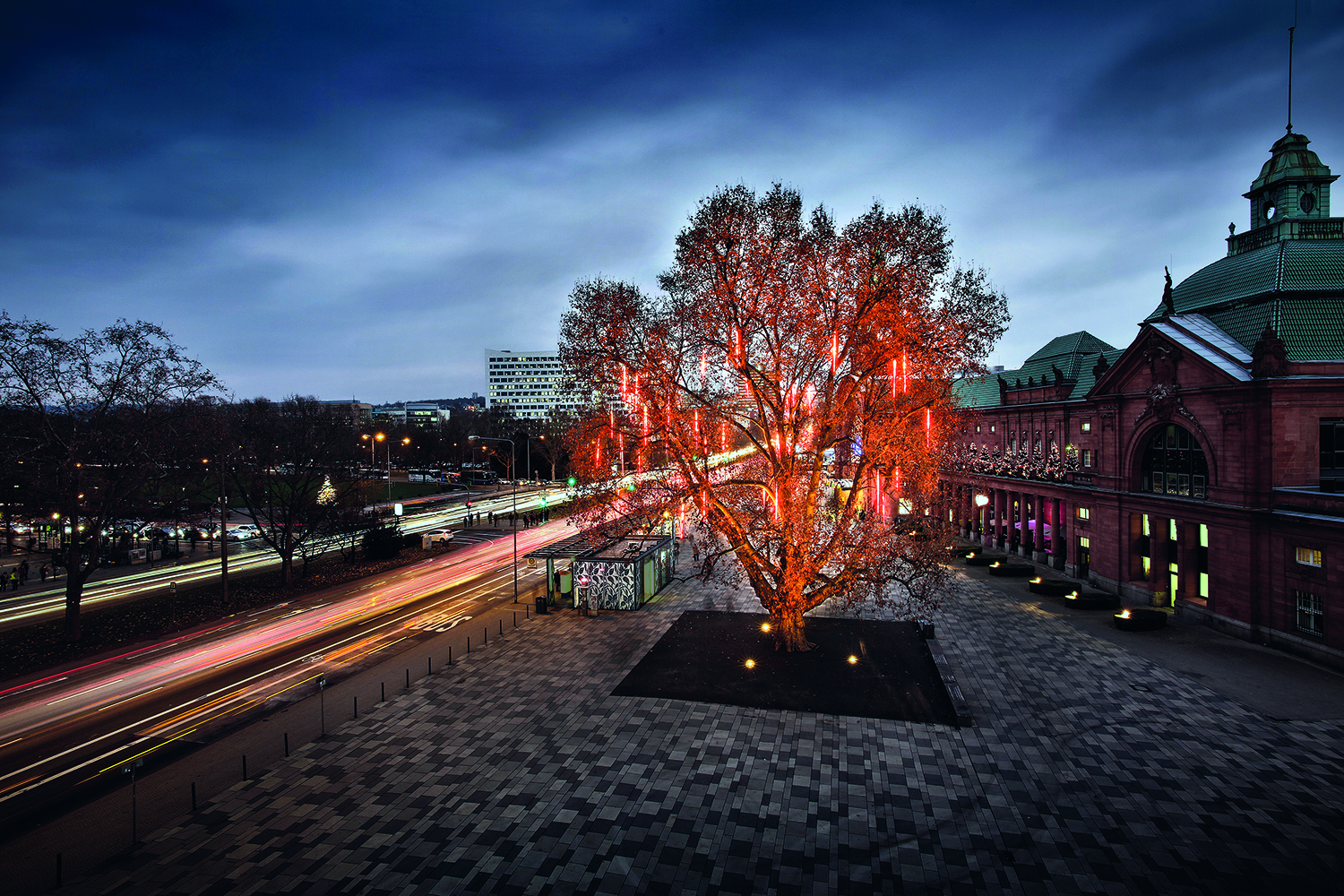 You can see a large tree with red light tubes hanging from its branches on a busy road at night