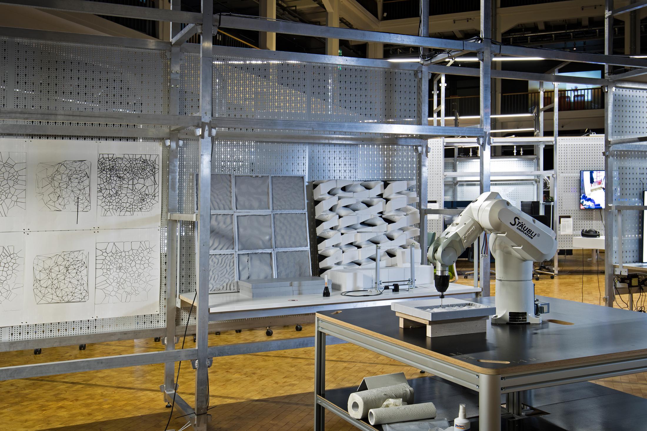 In the foreground is a robot arm on a wooden table, while in the background you can see various sculptures and drawings on a grid structure