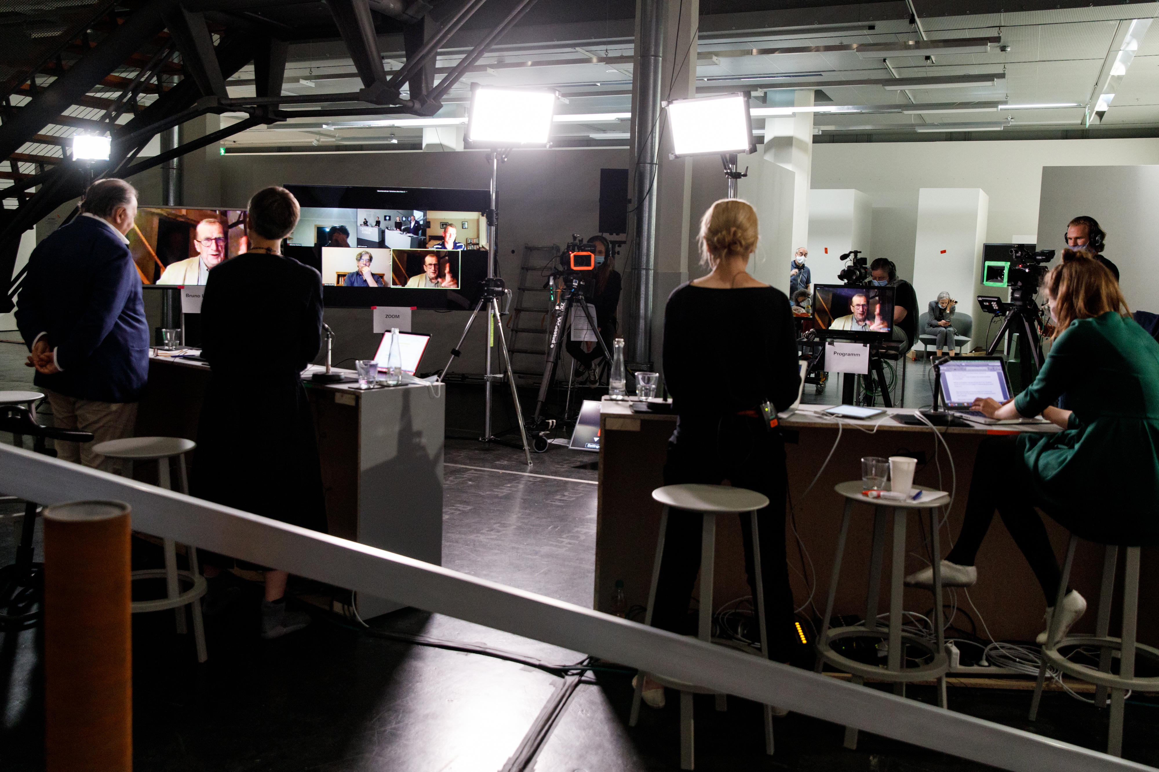 Peter Weibel and three of his colleagues stand at desks and are illuminated by spotlights while cameras are pointed at them. They look at large screens that show Bruno Latour and other streaming festival participants via video transmission.