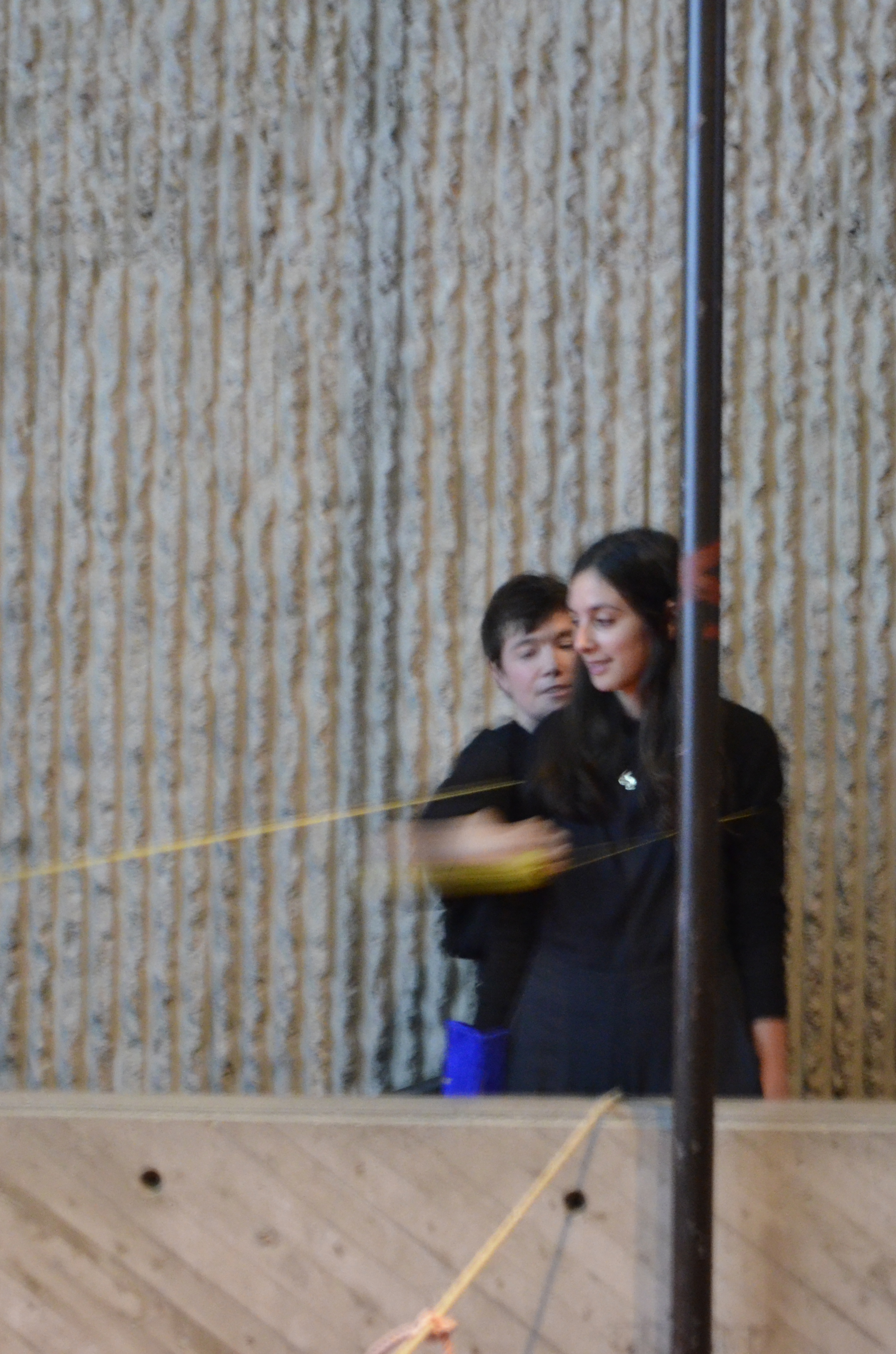 The photo shows two women standing diagonally behind each other. They wear dark clothes and stand in front of a grey wall. Different ropes can be seen schematically.