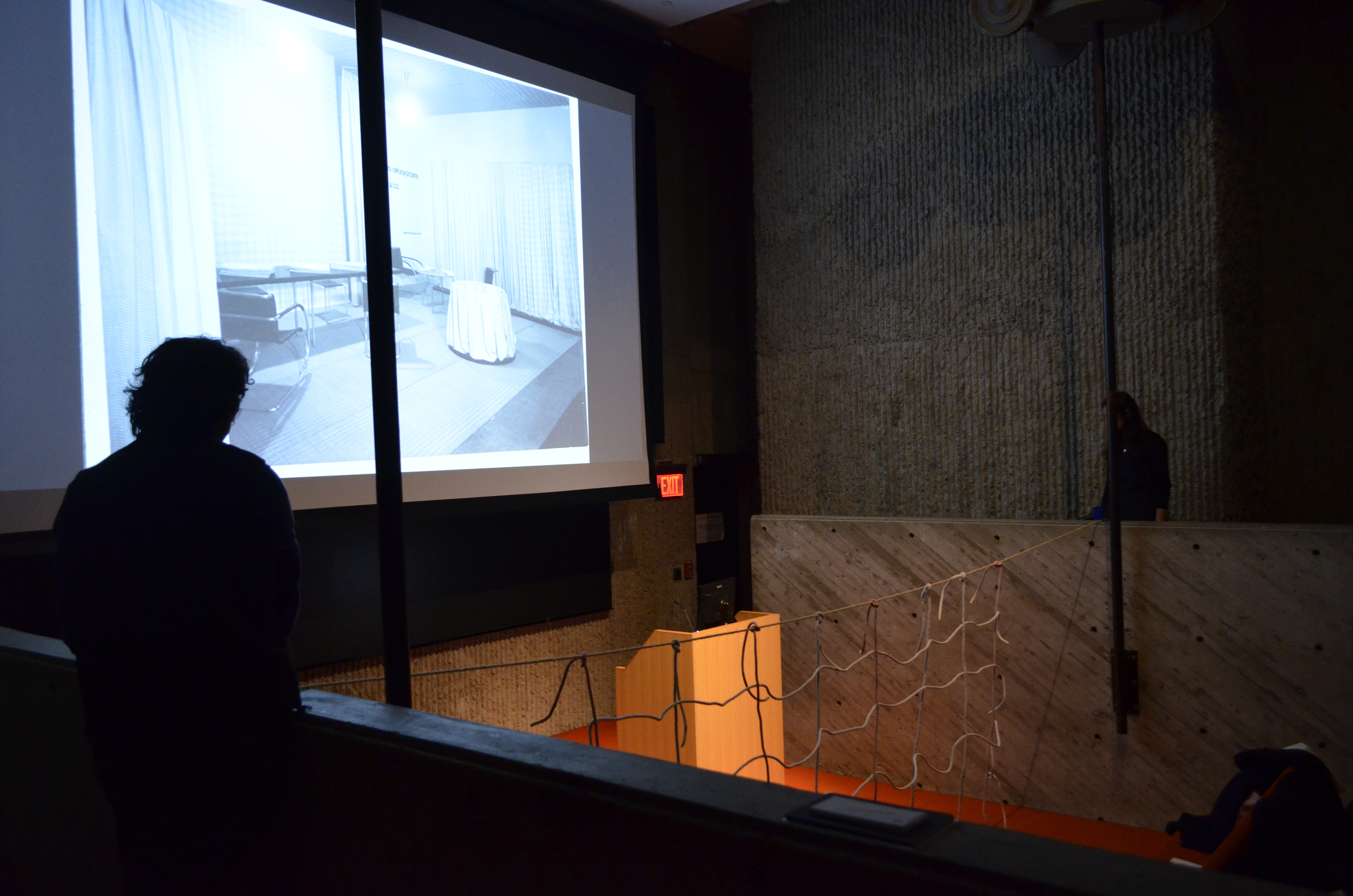 In the photo, the brutalist lecture hall is darkened while on the large screen an office-like room with curtains can be seen. In the middle hangs the net of ropes, which resembles a volleyball net.