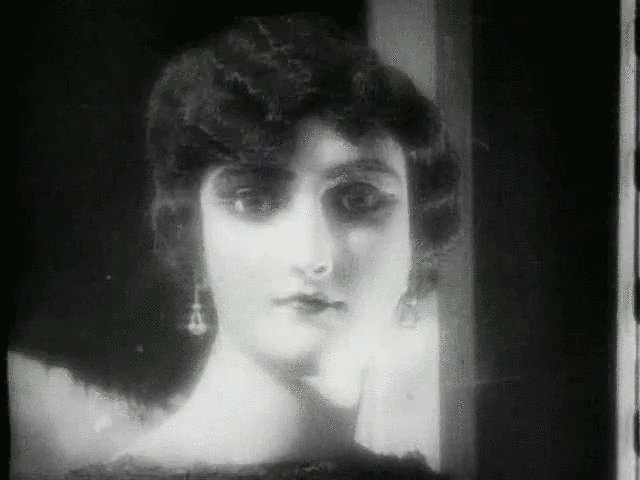 The screenshot shows a woman's profile in black and white. She looks into the camera from the front and looks almost like a doll.