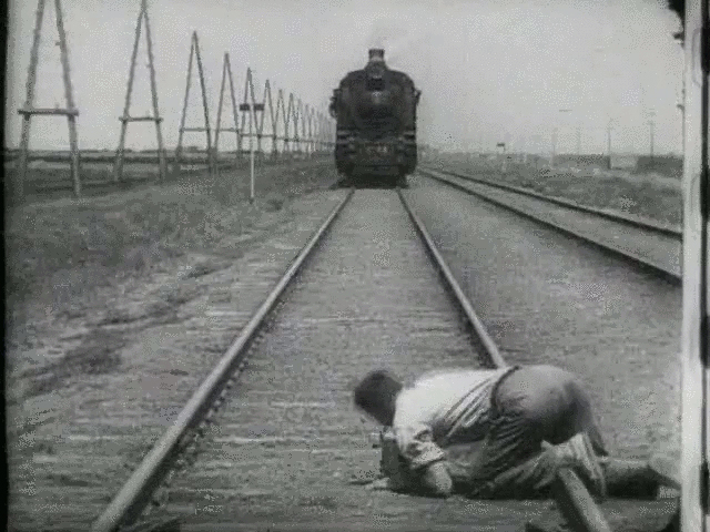 The black and white screenshot shows a track with a railway. A man lies crouching over a track and looks in the direction of the upcoming train.