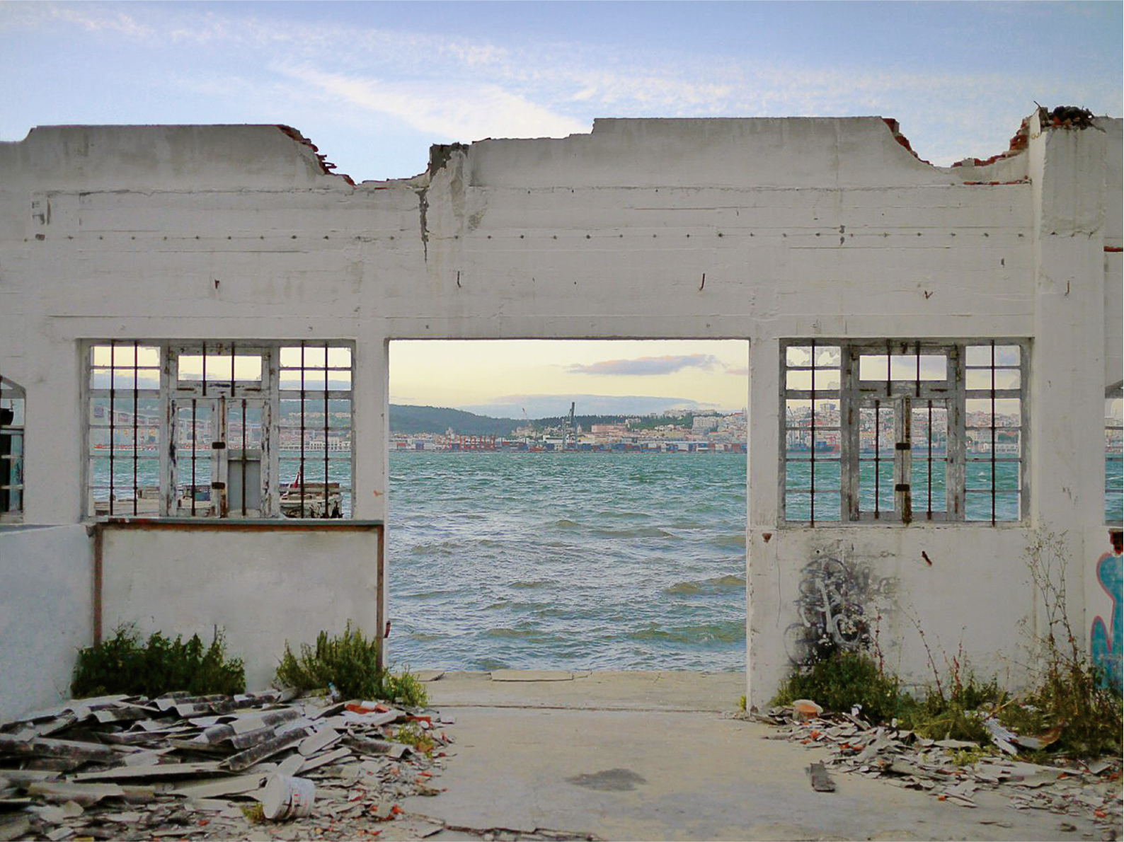 The picture shows a house ruin, through whose open doors and windows you can see the sea and a city behind it. 