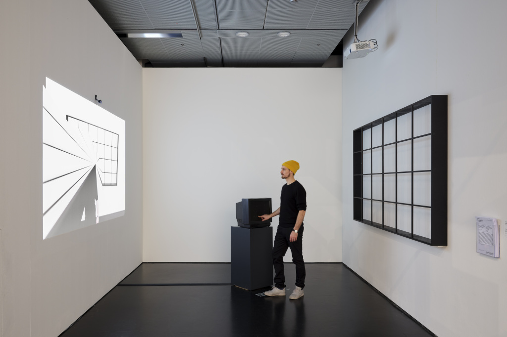 The photo shows a projection on the left and a rasterized image on the right wall. In the middle, a man in a cap holds his hand in a tube television.