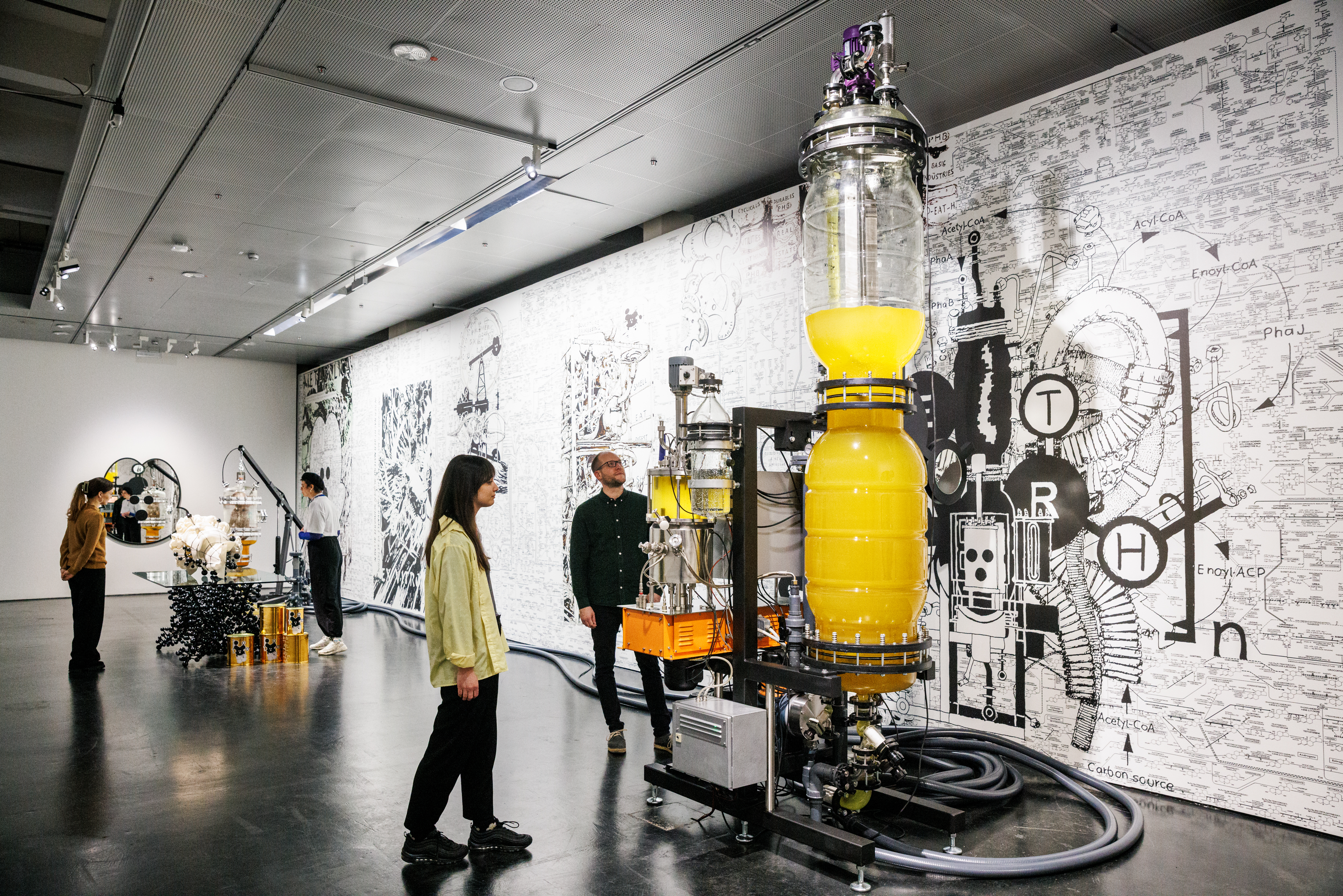 A ceiling-high, elongated glass vessel containing a yellow liquid can be seen. Several cables or hoses lead to smaller glass vessels that also contain liquid. Large hoses are lying on the floor. Two people are looking at this.