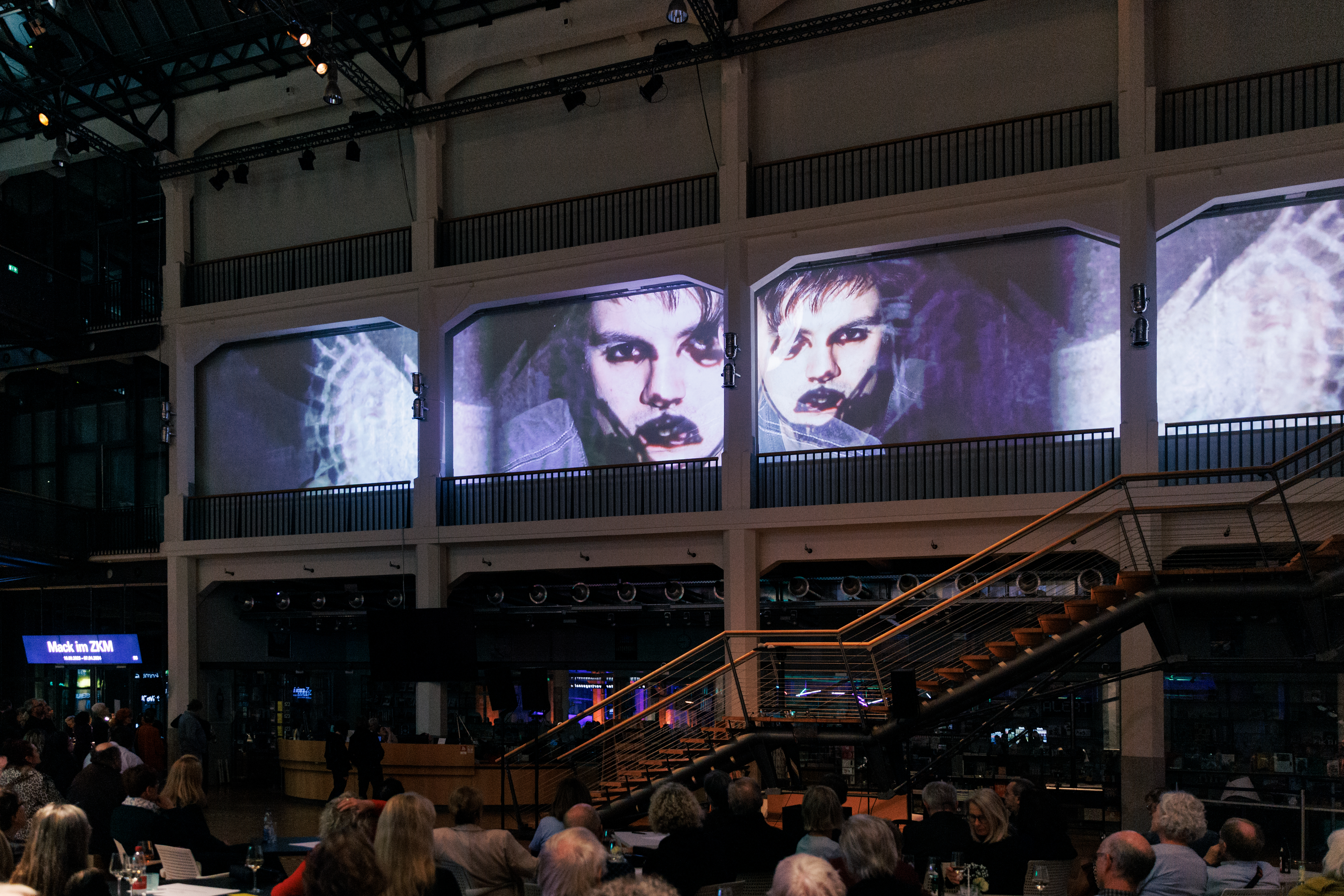 Das Foyer des ZKM | Karlsruhe ist voll mit Besuchern, die alle hoch an die Wand schauen, auf die ein Video projiziert wird. Auf dem Video zu sehen sind zwei Gesichter.