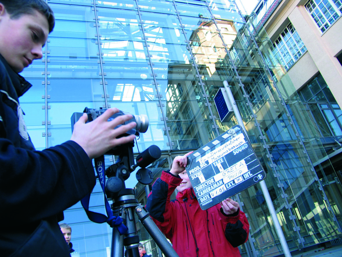 Someone is filming with a camera that is standing on a tripod. Somebody else is holding the clapper board in front of the camera.