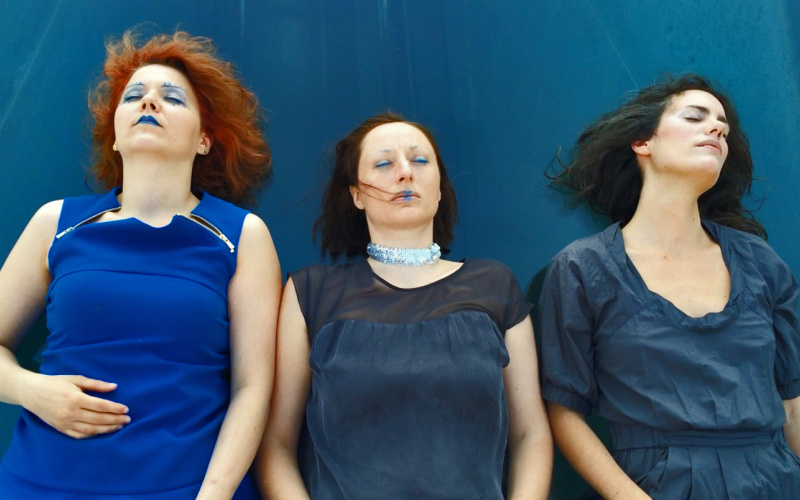 Three women dressed in blue and wearing blue makeup, lying on the blue tarp