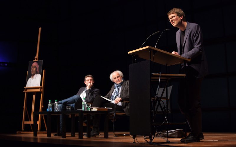 Links: An easel with a picture of a man with a turban. Middle: Two men sitting on chairs. Right: A man standing at a podium and addresses the audience.