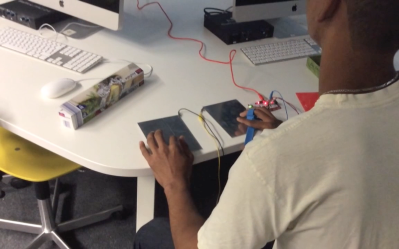 A boy is sitting in front of a computer and on the table in front of him there are two squares, a diy controller, by which he can control the videogame.