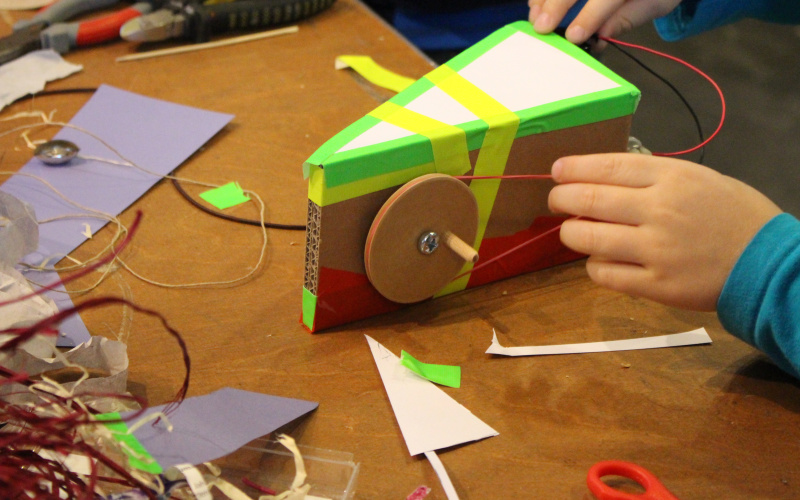 A kid works on a little automated construction, that it has equipped with a wheel, that is beeing rotated by a rubberband and a small motor.