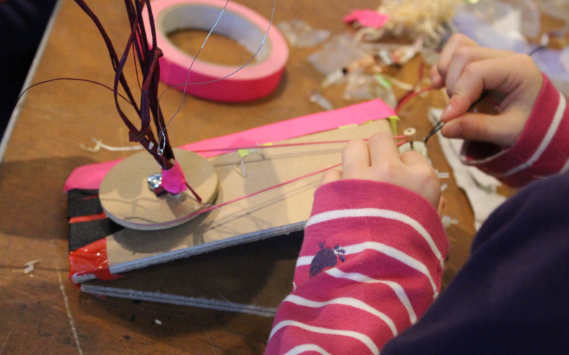 A kid works on a little automated construction, that it has equipped with a wheel, that is going to be rotated by a rubberband and a small motor.