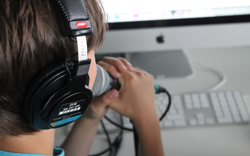 A boy is speaking into a microphone that he is holding infront of his mouth. Meanwile he looks into the computer on the table at which he is sitting.