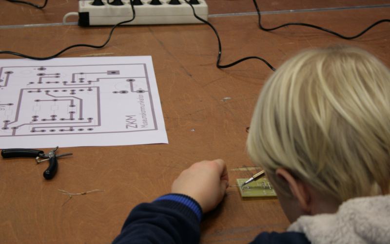 A boy is working on a circuit board with a soldering iron, out of which he is buil