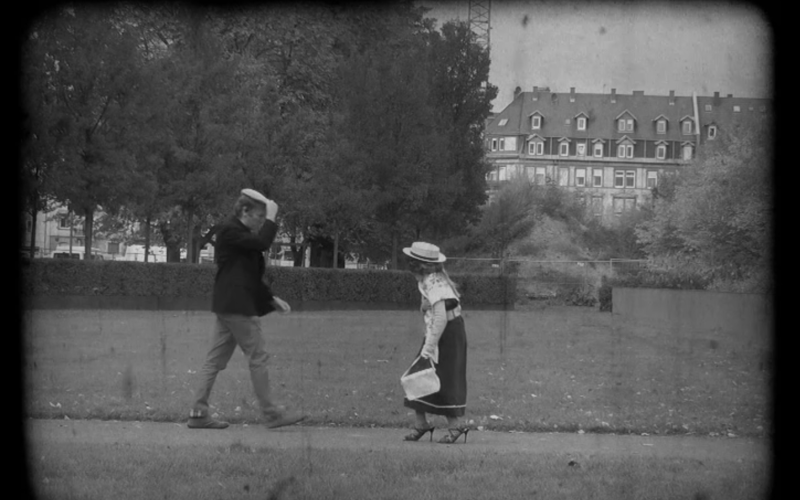 Two pedestrians meet on the street, the man is lifting his heat as to greet the Woman, that is aproaching. It is a still from a black-and-white movie made in the workshop "the little rascals". 