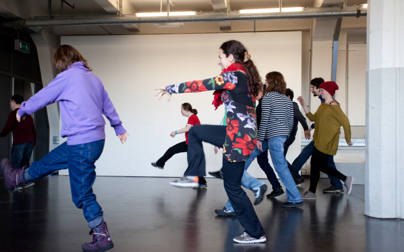 A group of young people is dancing exuberantly whilst throwing the legs in the air and laughing happily.