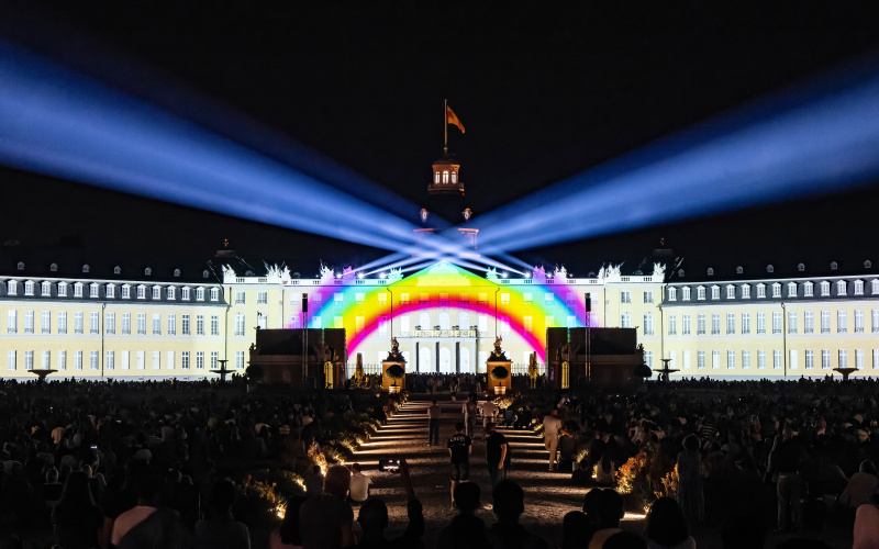 Zu sehen ist die Fasade des Karlsruher Schlosses. Darauf projeziert ist ein Regenbogen mit Scheinwerfern, die in den schwarzen Himmel leuchten.