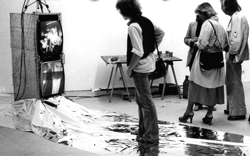 A man is standing on a silver foil and looks at a video installation.