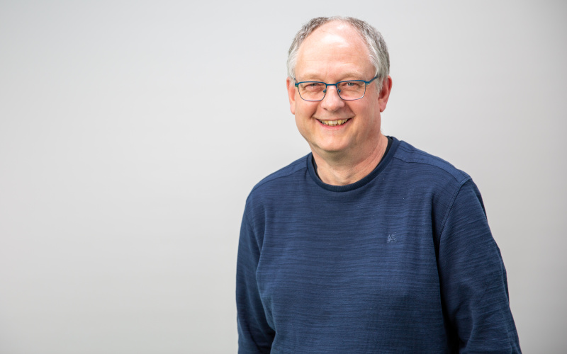 stehender Mann mit grünem Pullover, grauen Haaren und Brille