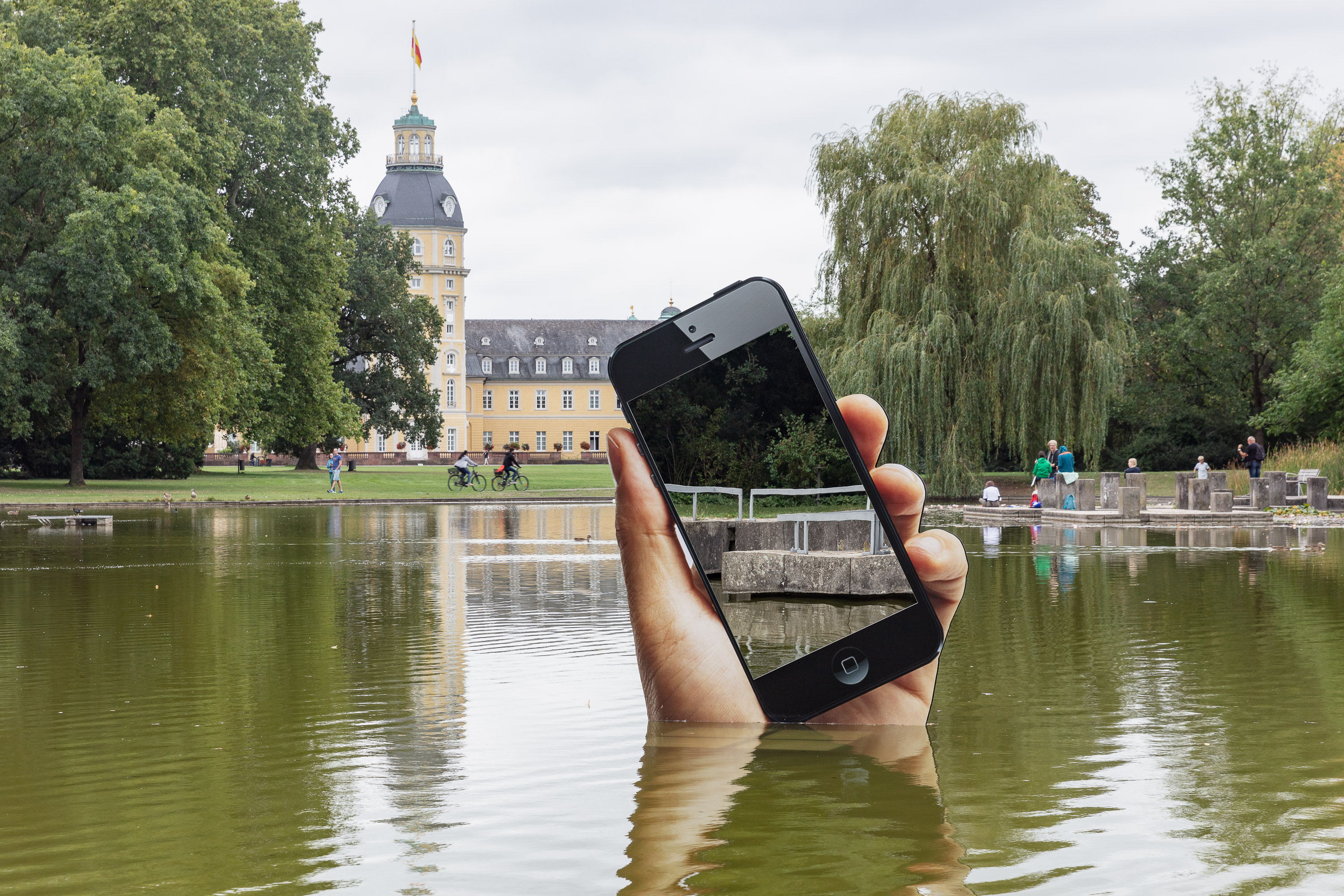 You can see the shore of a lake. A very large hand with a smartphone is sticking out of the lake. The display of the smartphone is a mirror in which the viewers can see themselves on the shore.