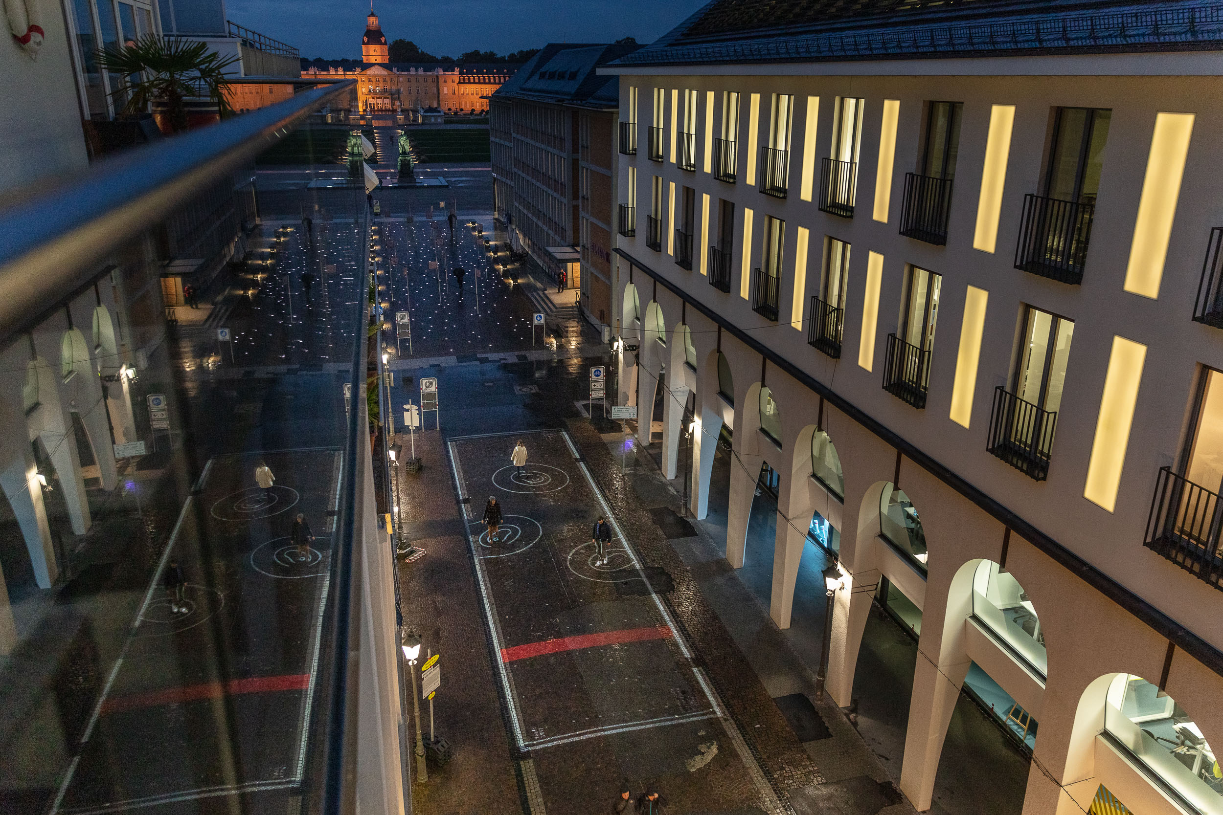 You can see the view of a street from above. On the floor are spiral circles of light. In the background the castle of Karlsruhe is visible. 