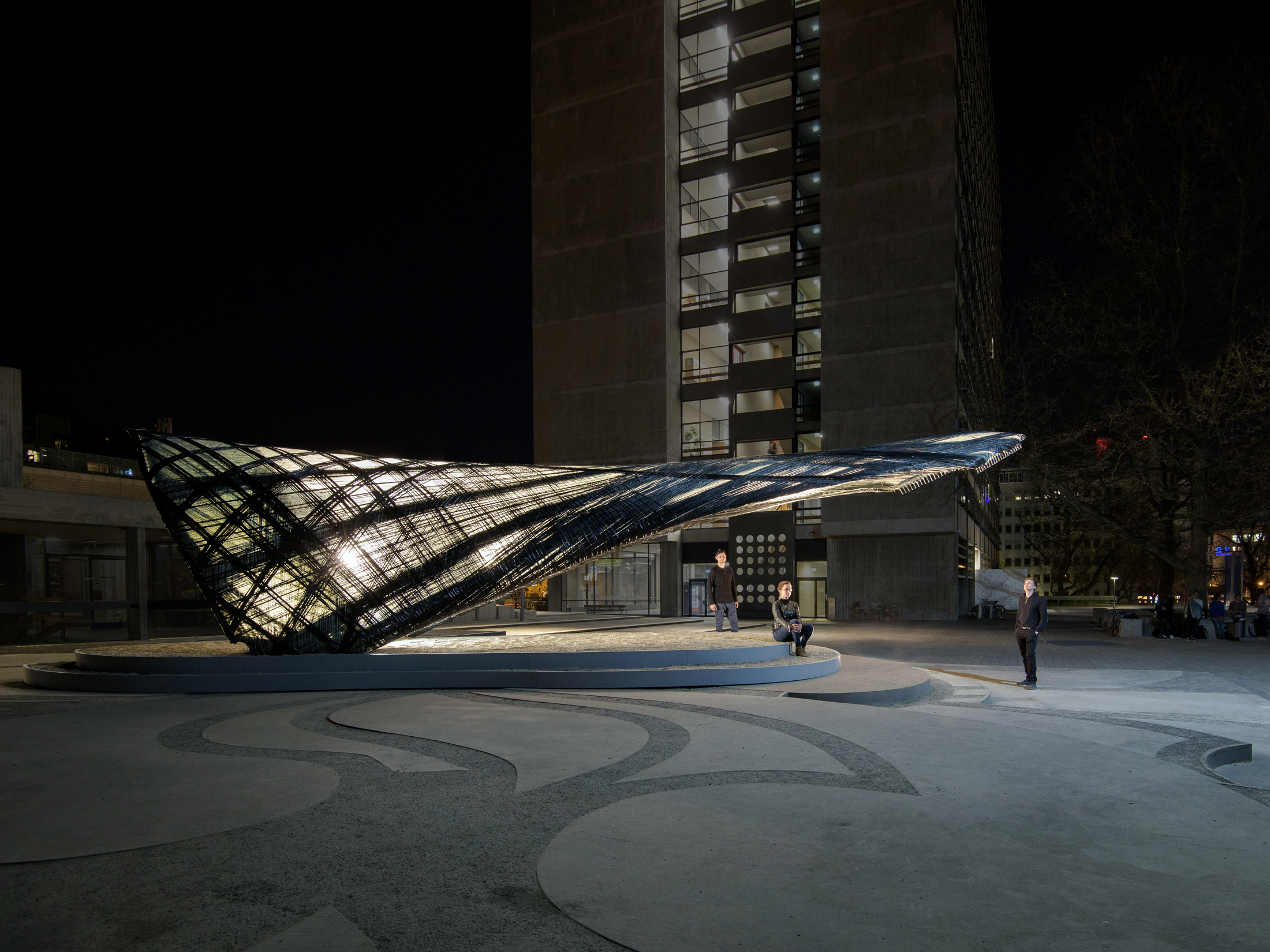 The picture shows a seated woman and two men standing around an illuminated research pavilion made of glass and carbon fibres