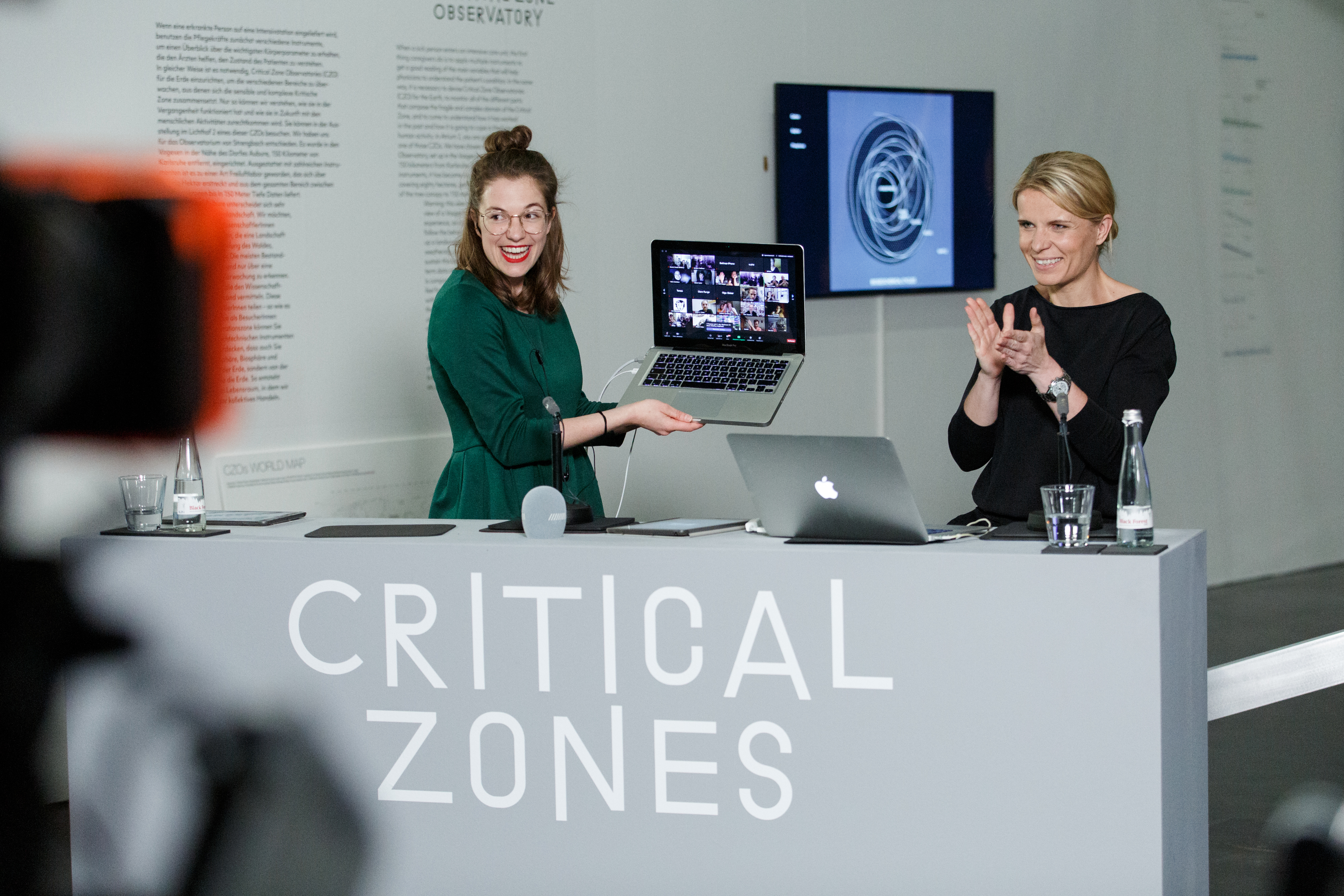 Two women stand behind a desk. One woman is holding an open laptop, beaming with joy.