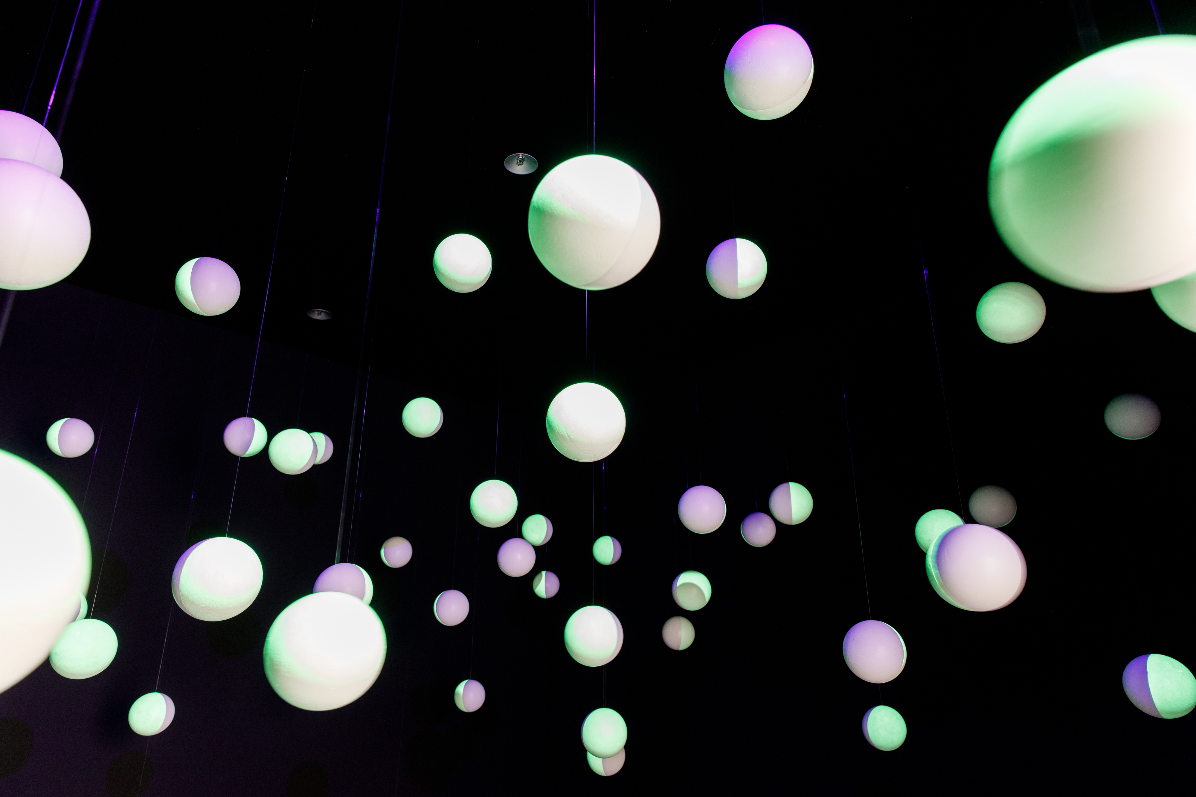 White polystyrene balls hang in a black room, the balls are painted with fluorescent paint and are illuminated.