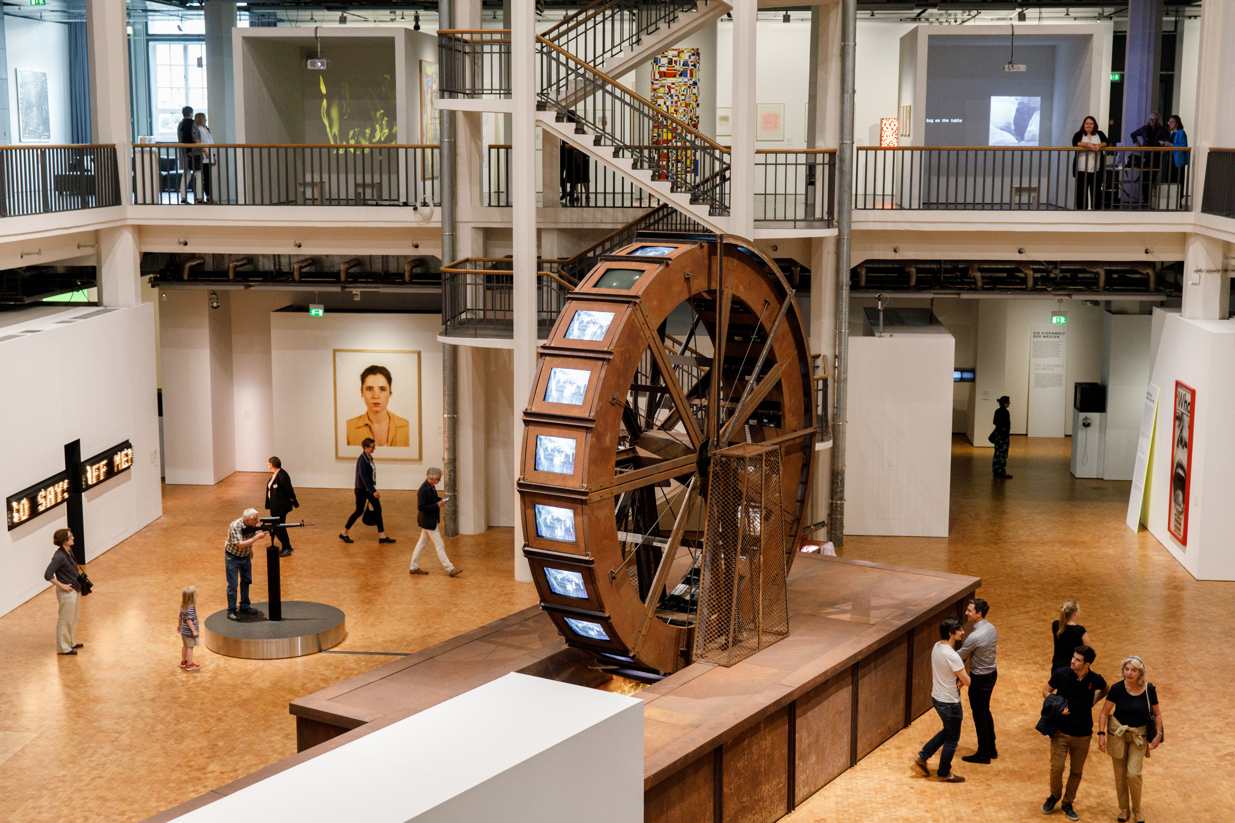 A huge metal mill wheel stands in an exhibition room. At the mill wheel there are tube screens on which videos of water are played.