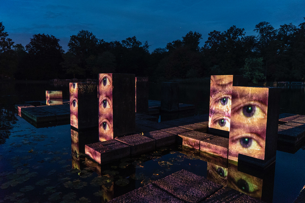 Stelae inside a lake in a park. It is late evening. Eyes are mapped onto the stelae, which look openly into the area