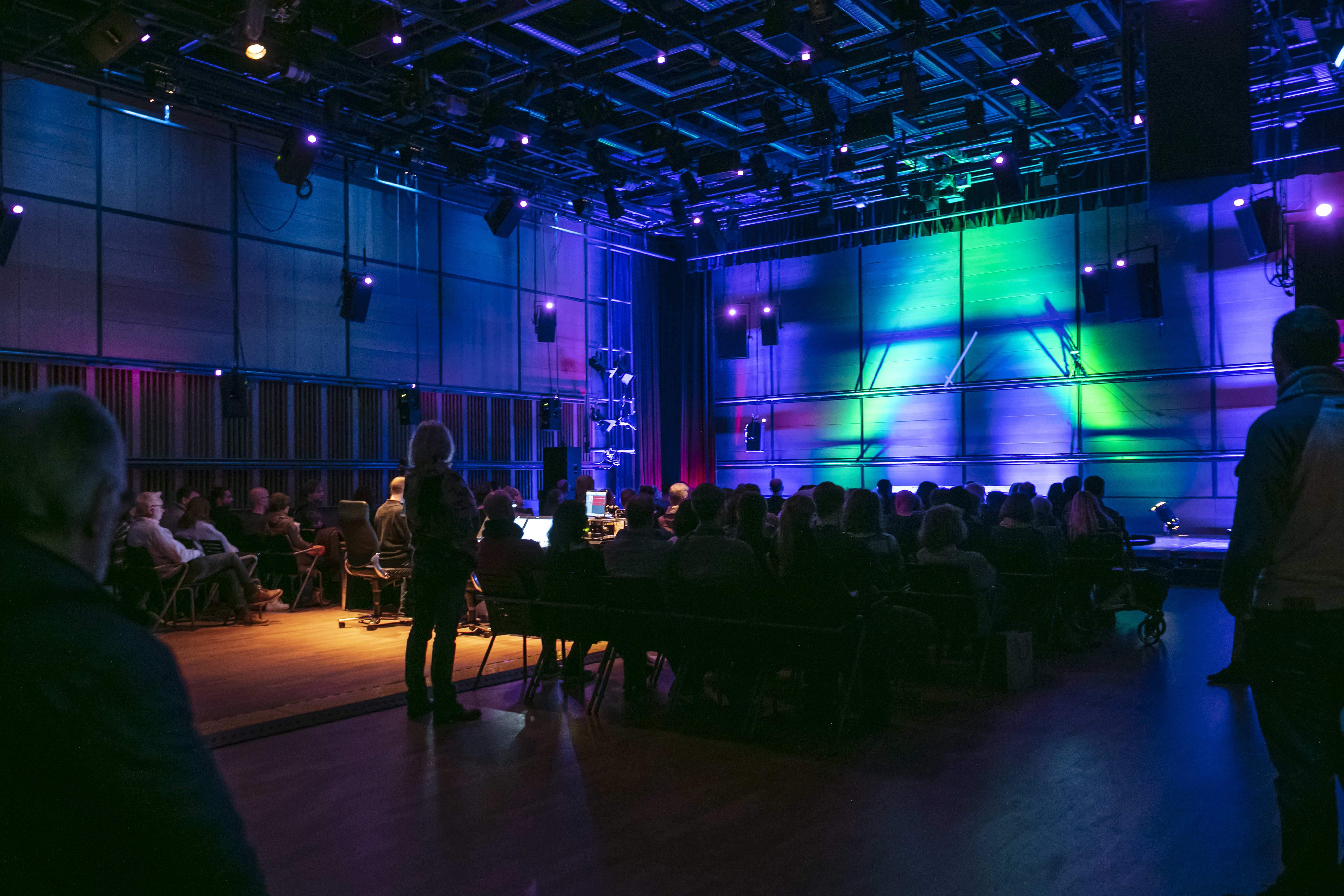 Ein abgedunkelter Raum mit sitzenden Menschen die auf eine Leinwand mit blauen und gelben Lichtstrahlen schauen.