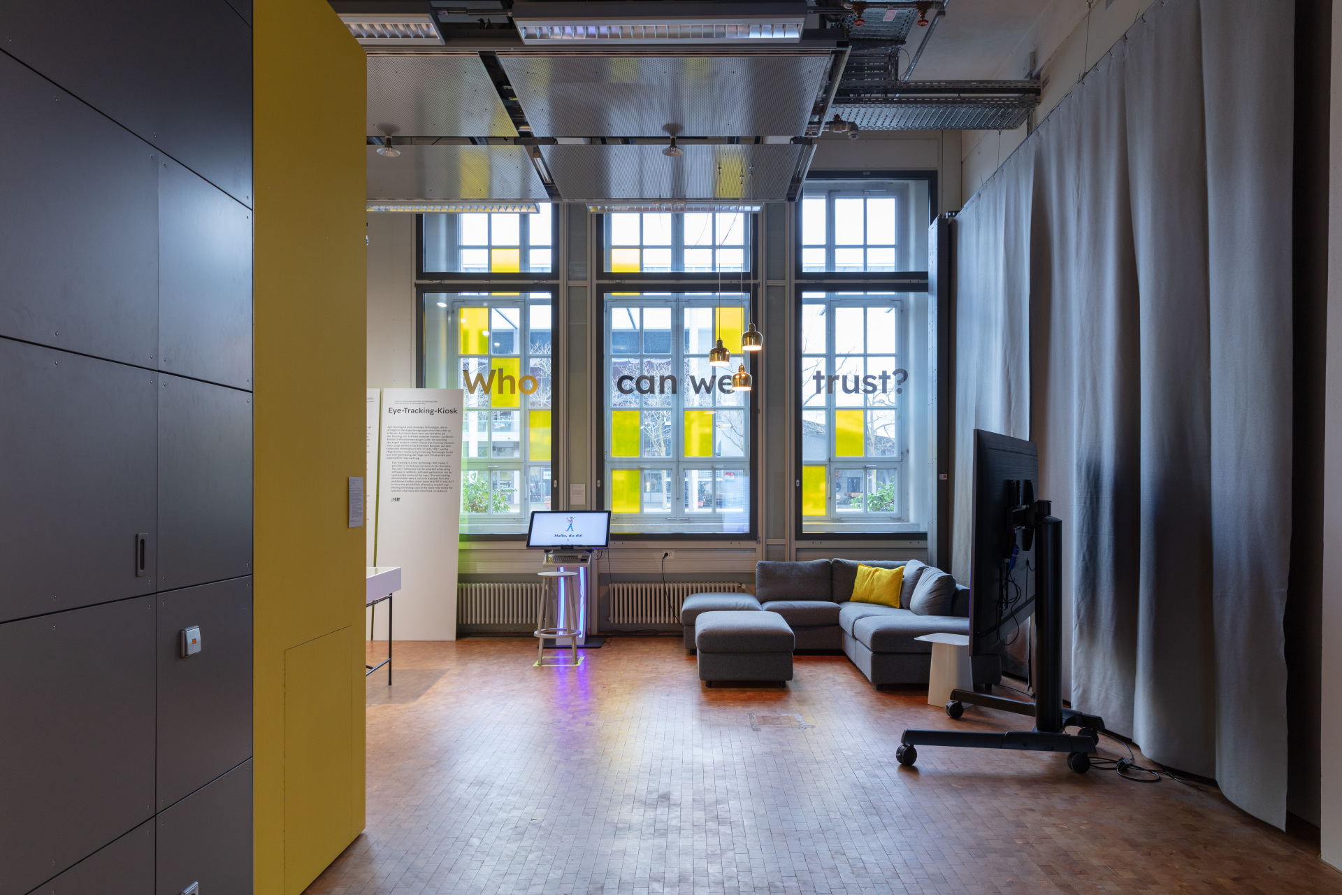 View into the exhibition of a yellow wall, a couch and the work "eye-tracking kiosk" consisting of a screen and a stool stealing away from it