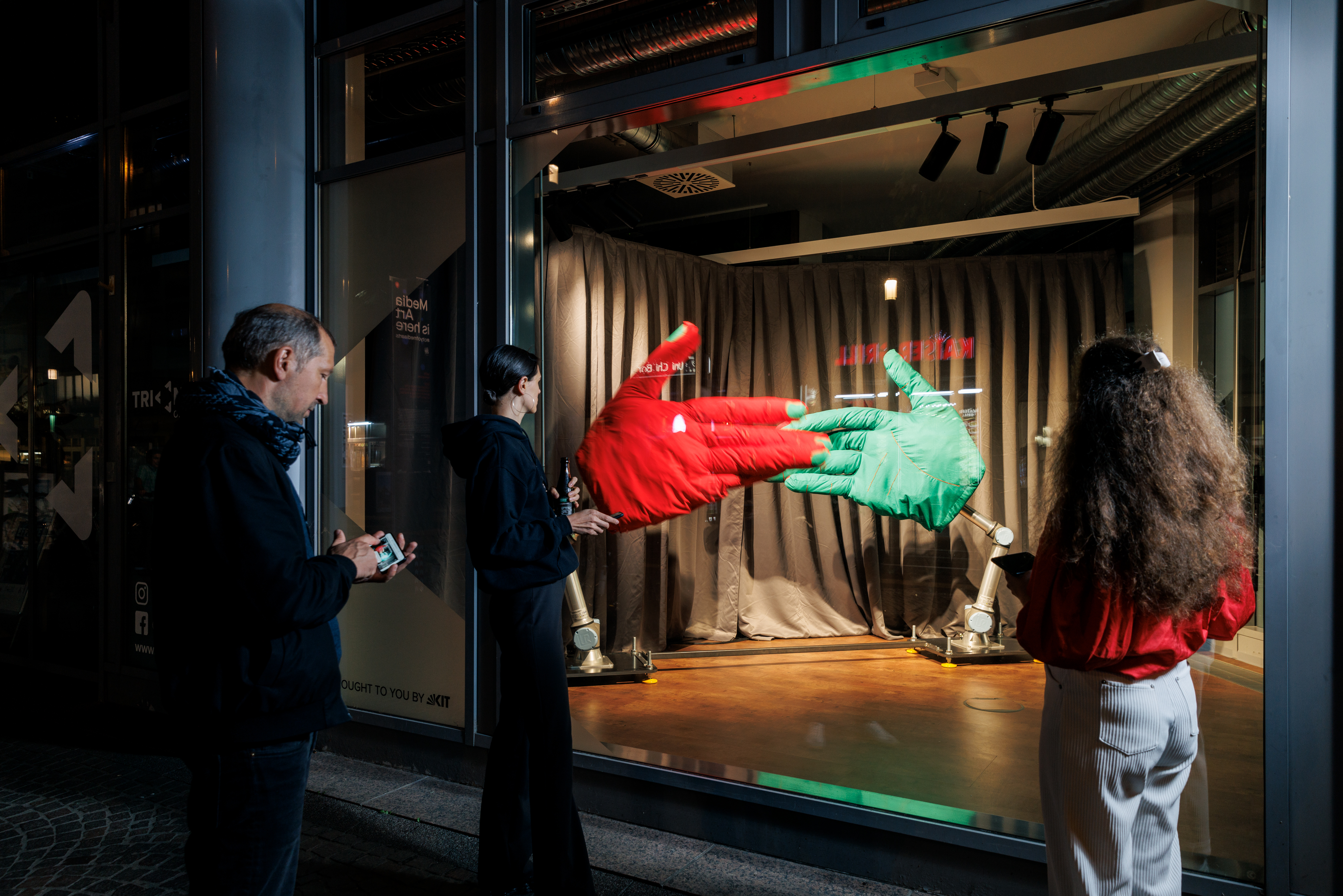 Auf dem Bild ist die Instalation »Handshake«,  AATB (Andrea Anner, Thibault Brevet), im Triangel am Kronenplatz zu sehen. Eine große ror und große grüne Hand sind am Boden fixiert und schlagen sich gegenseitig ein. Mehrere Menschen stehen vor der Scheibe.