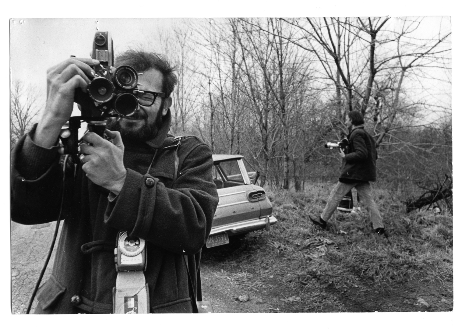 A black and white image of a man who also takes a photo. In the background a car and another man running in the picture