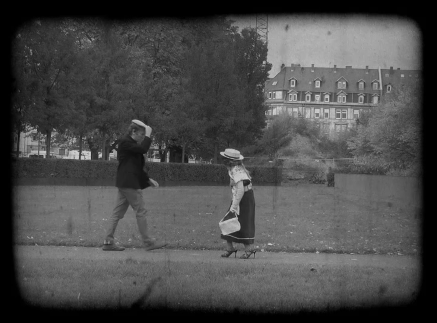 Two pedestrians meet on the street, the man is lifting his heat as to greet the Woman, that is aproaching. It is a still from a black-and-white movie made in the workshop "the little rascals". 