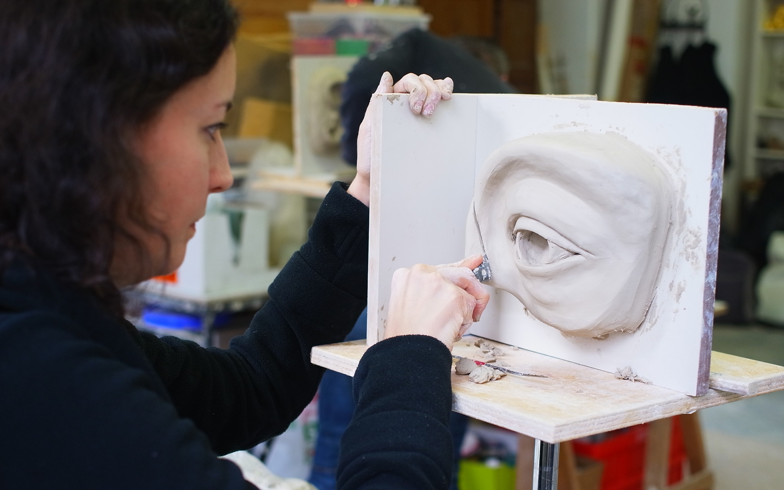 A woman is working on a sculpture of an eye area out of clay. She is cutting into the clay with a knife.