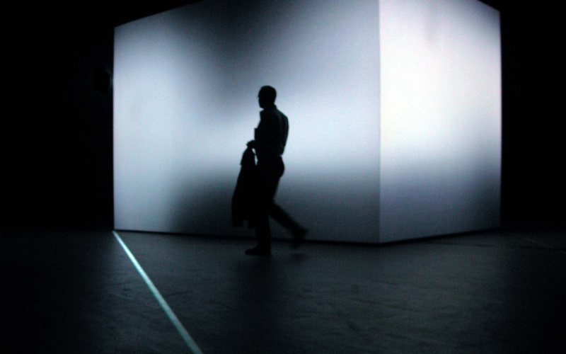 In einem völlig dunklen Raum steht ein 3,5 Meter hoher Würfel, dessen Form durch eine Lichtprojektion verfremdet wird. Davor der Schatten eines Mannes..