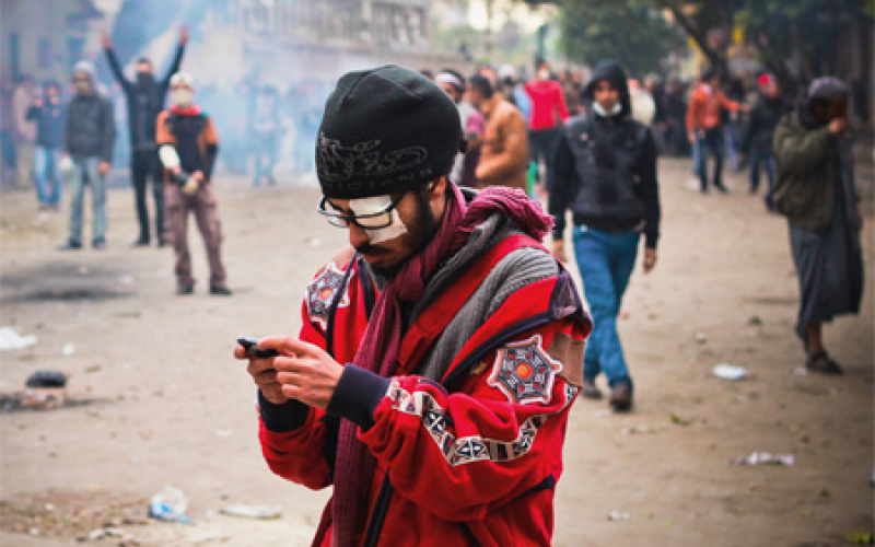 Ein Mann in roter Jacke und schwarzer Mütze. Sein linkes Auge ist verbunden. Er trägt eine Brille und schaut auf etwas, was er in seinen Händen hält. Im Hintergrund eine Vielzahl von Menschen.