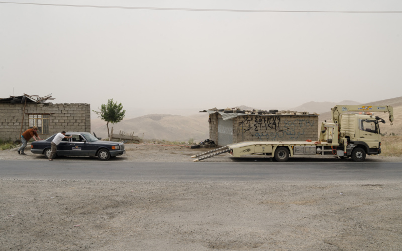 Two men are pushing a mercedes towards an tow truck. Behind them are ruinous buildings and a gaunt hilly landscape