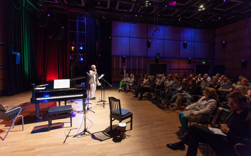 A man standing on a stage. There are several chairs and a piano