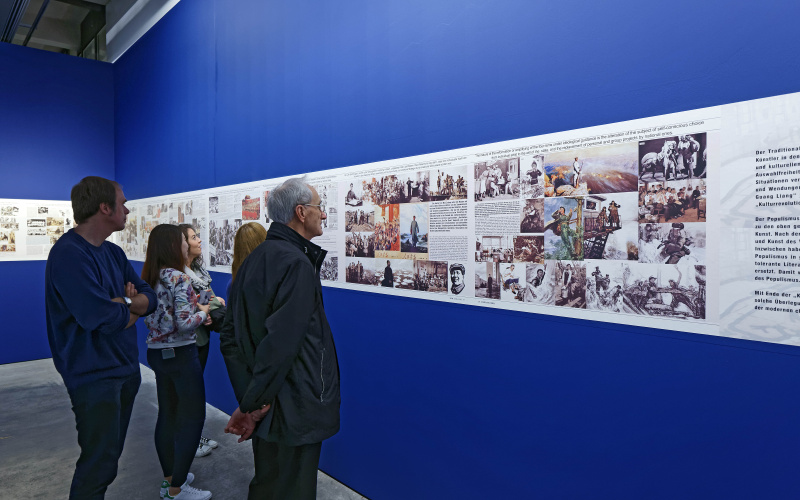 Five people look at pictures in an exhibition, which are hanging on a blue background