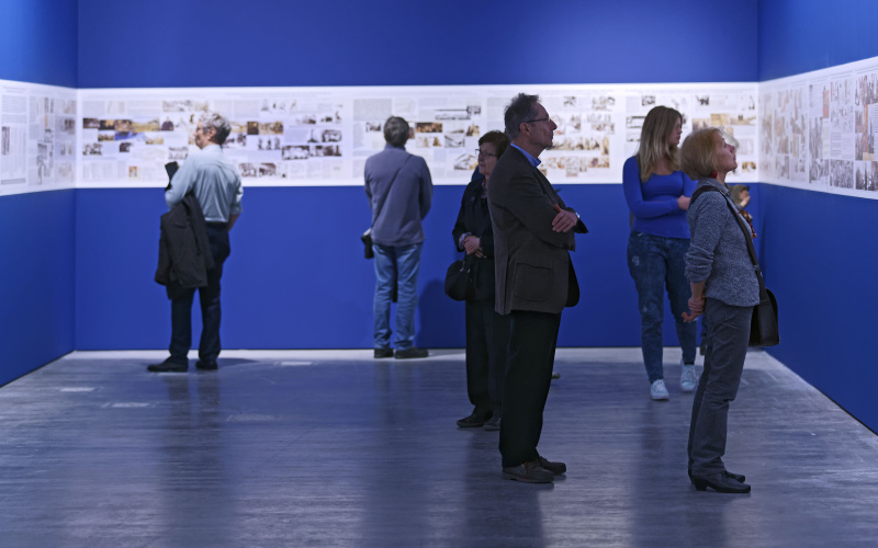 Menschen sehen sich Bilder der Ausstellung an, die auf blauem Hintergrund ausgestellt sind