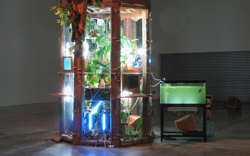 In a type of greenhouse, that is illuminated by spotlights from the inside, are lots of plants and computers. Next to it is an aquarium that is connected to the greenhouse by tubes. At the Rim of the glasshouse there is a wooden figurehead.