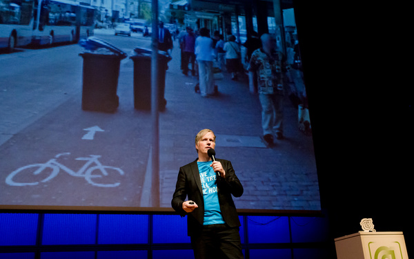 A man on stage. In the background, on the screen, a  bicycle path